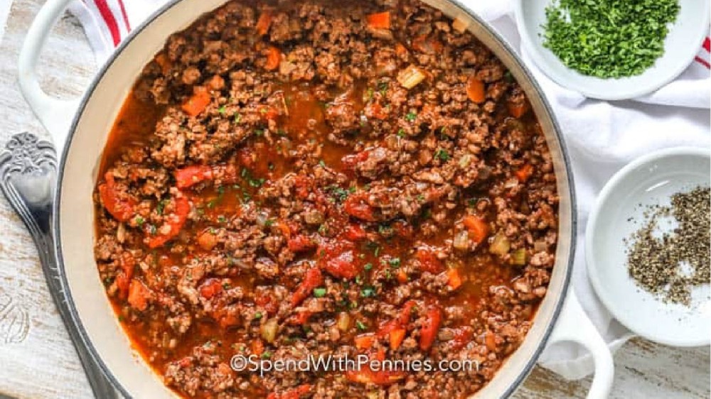 An enamed cast iron Dutch oven filled with a hearty bolognese sauce with pepper and herbs on the side. 