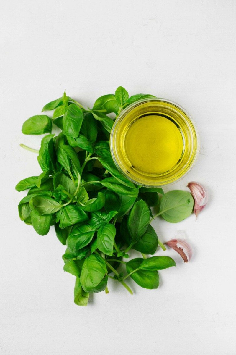 A jar of olive oil, basil leaves, and cloves of garlic have been laid out onto a white surface.