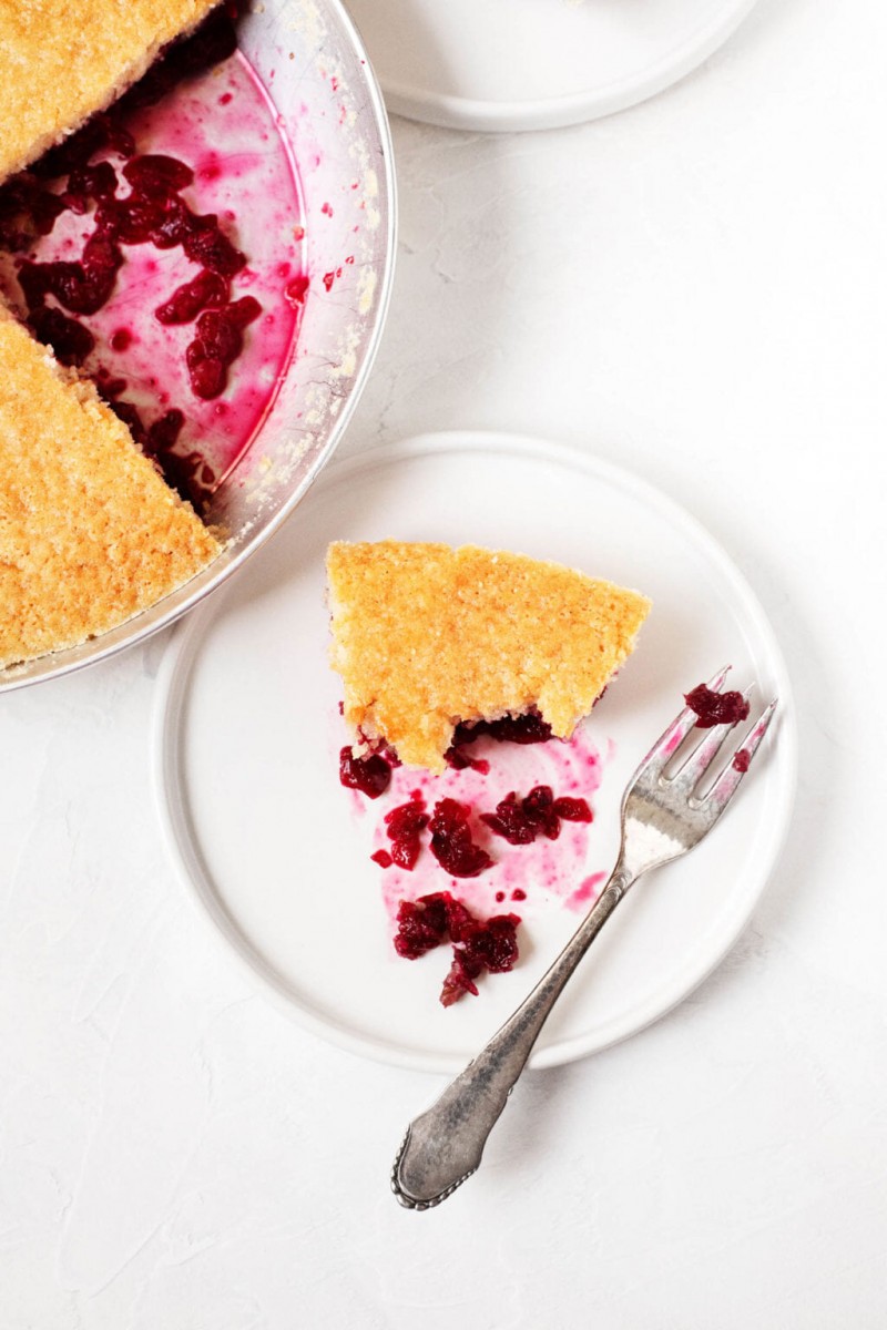 A slice of cake with cranberries on the bottom rests on a dessert plate with a fork and a pie plate nearby.