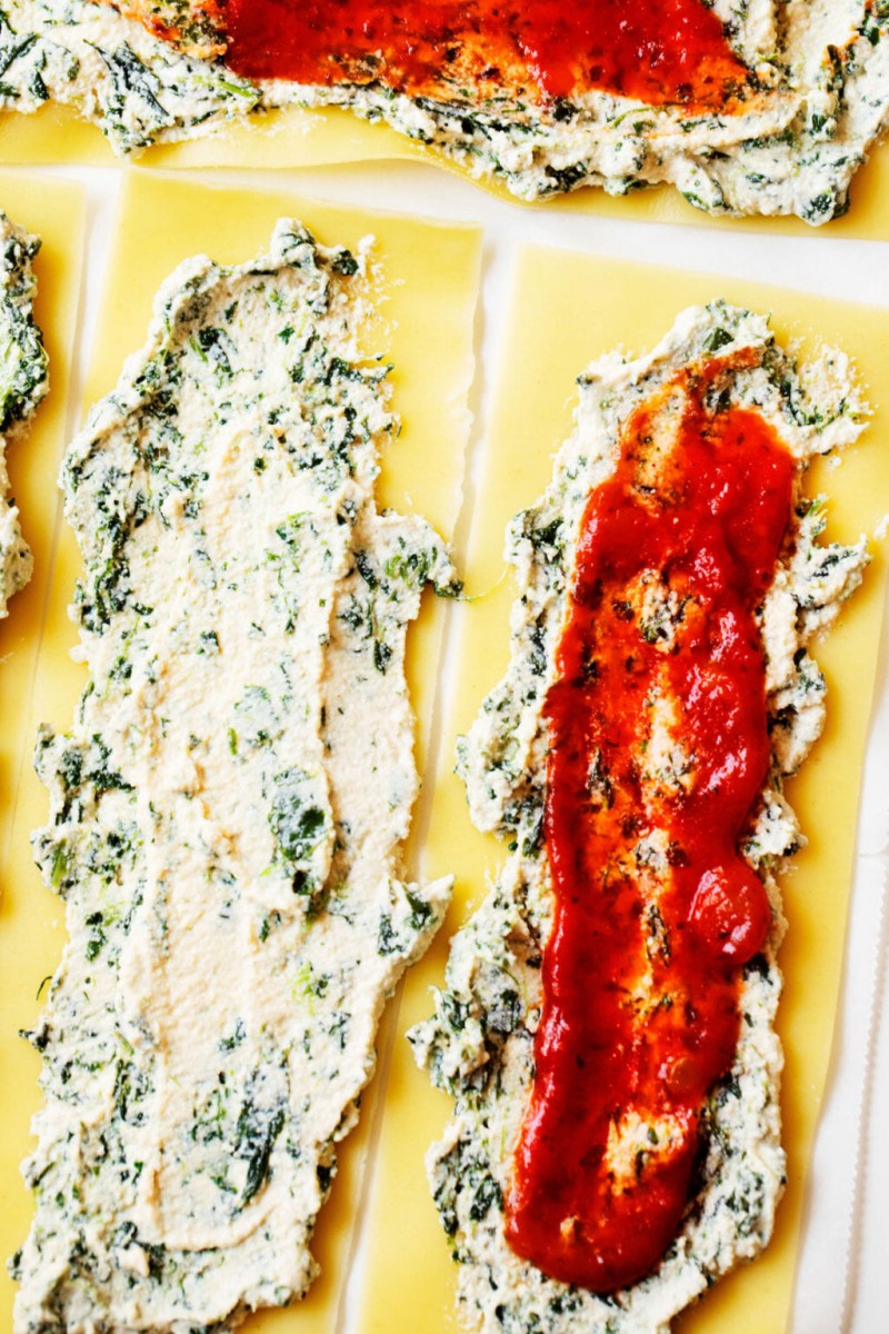 A few lasagna sheets are laid out on a work surface. Each is covered with plant-based ricotta, spinach, and marinara sauce.