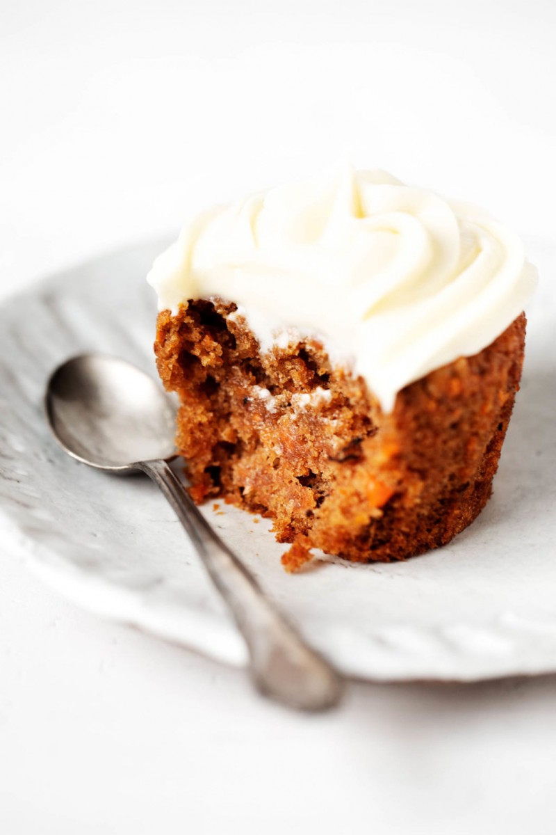 A vA vegan baked good with frosting has been bitten into. It's resting, along with a spoon, on a fluted white ceramic plate.