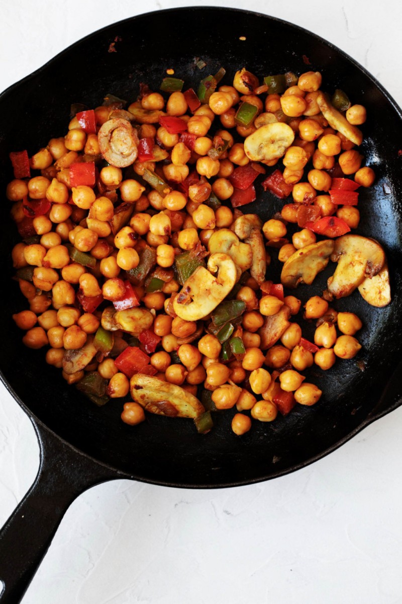 A black, cast iron skillet holds a mixture of sautéed vegetables and garbanzo beans.