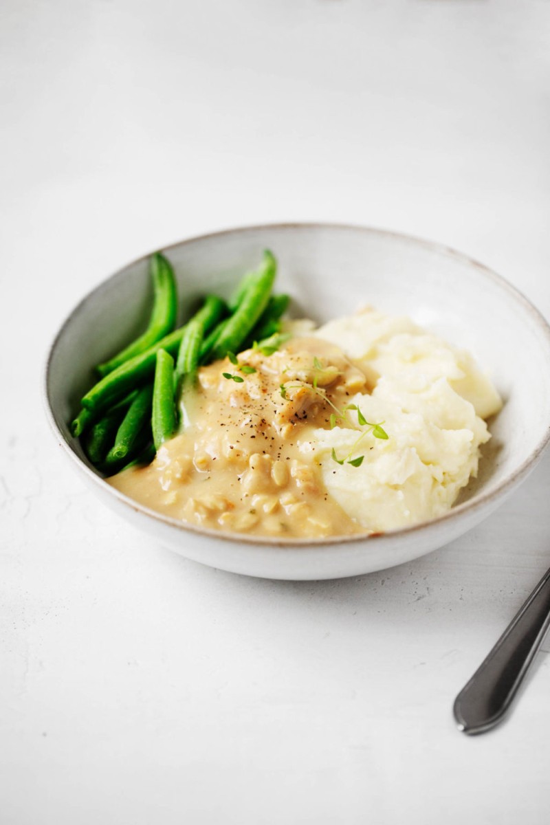 An angled photograph of a serving of plant based ingredients, including green beans and soy tempeh with gravy, garnished with herbs.