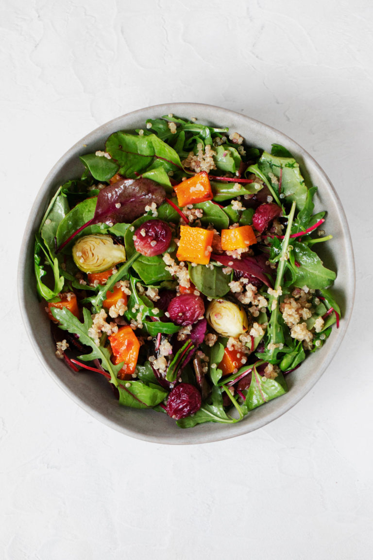 A round, white bowl has been filled with a vibrant roasted butternut squash salad that includes a lot of bright, leafy greens.