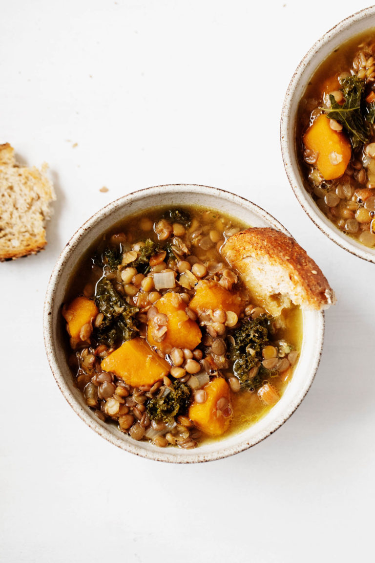 Two bowls of butternut kale lentil soup are laid on a white surface. A piece of toast rests nearby.