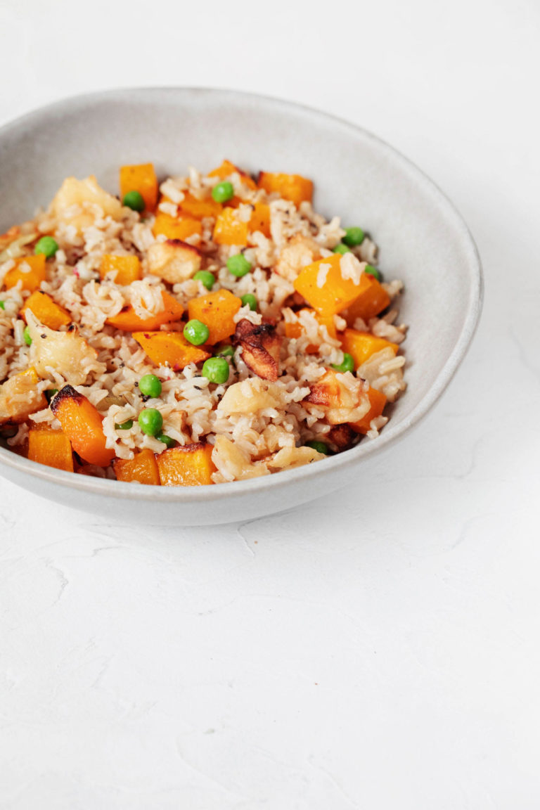 A white, ceramic bowl holds a mixture of roasted butternut squash, rice, and peas.