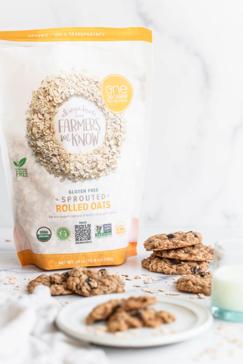 A few cookies are resting on a white surface. A bag of whole grains is pictured to the left and back of the cookies.