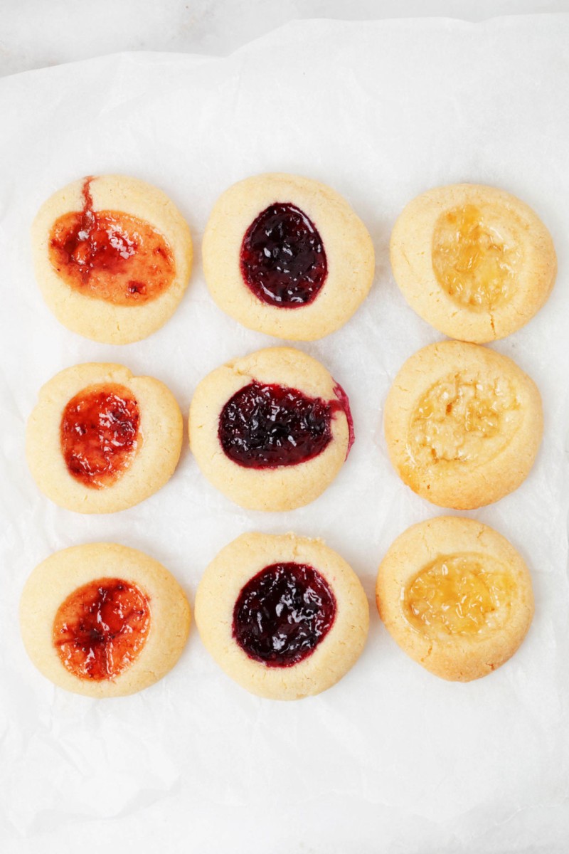 Three rows of thumbprint cookies, each filled with a different colored jam, are resting on a parchment lined baking sheet.