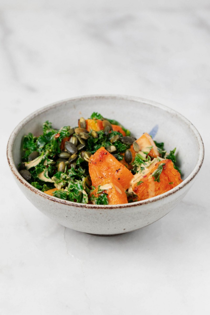 A white and grey ceramic bowl has been filled with steamed and roasted vegetables, along with toasted pepitas. It rests on a marble surface.