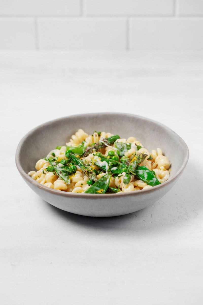 A white and gray ceramic bowl holds a dish of grains and bright green produce. It's topped with herbs and resting on a white surface.