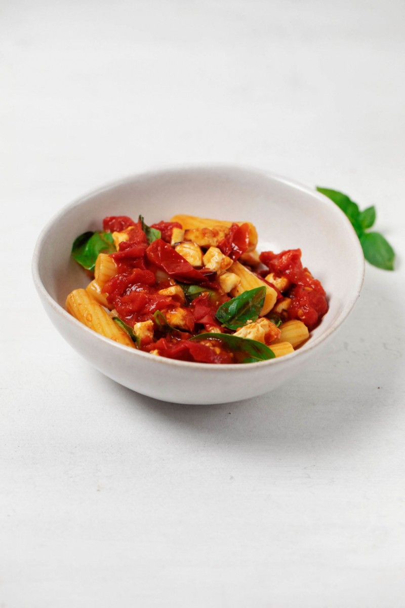 A round, white dish holds tomatoes, tube shaped pasta, and herbs.