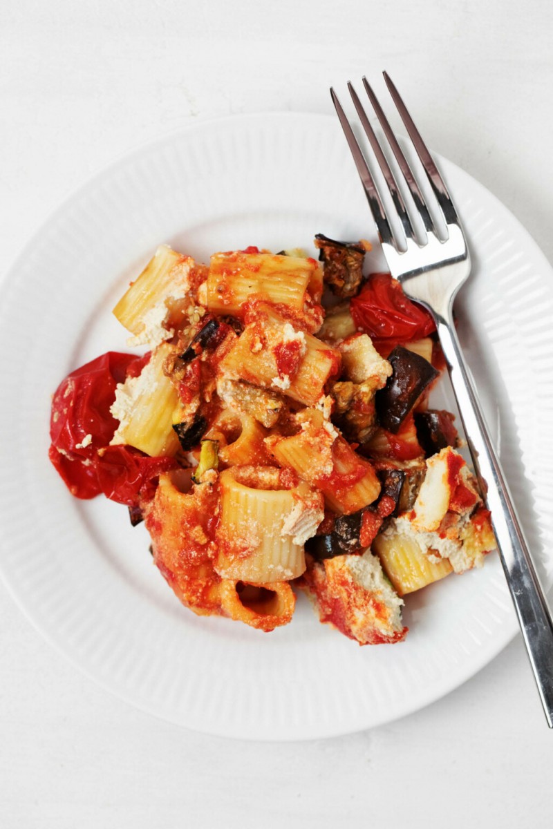 A vegan pasta bake has been portioned onto a round, white plate. A silver fork rests on the plate.