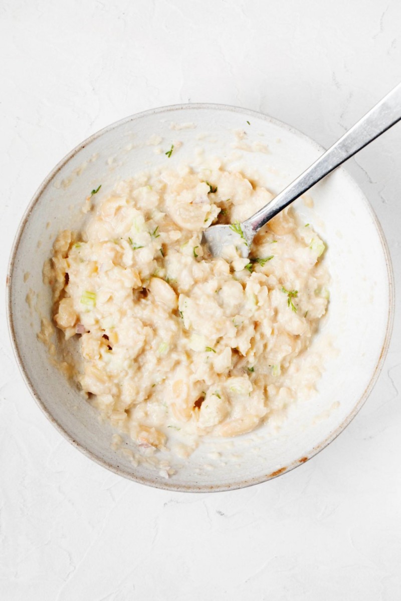 A smashed white bean salad is held in a white, ceramic bowl, resting on a white surface.