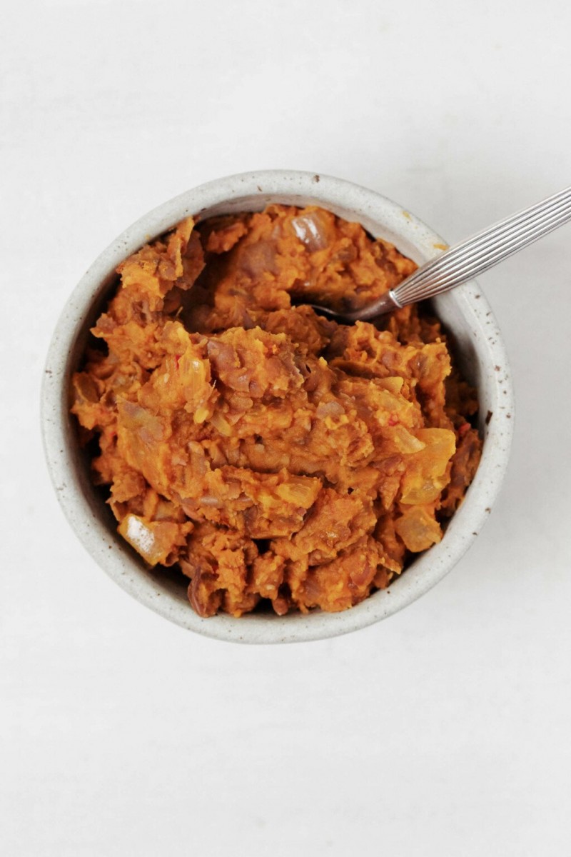 A small, white ceramic bowl has been filled with a brown mashed bean dip. It rests on a white surface.