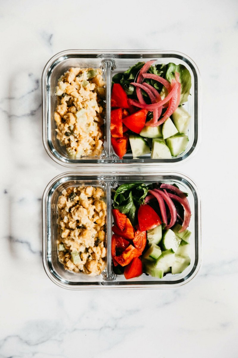 Two divided Pyrex containers have each been filled with smashed chickpea salad and vegetables. They rest on white marble.