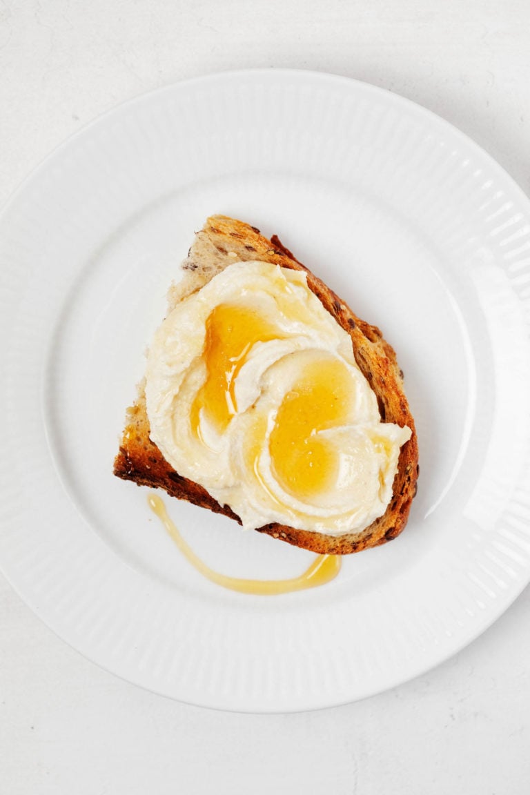 An overhead image of a round, white rimmed plate, which holds a slice of toast with vegan ricotta and agave drizzle on top.