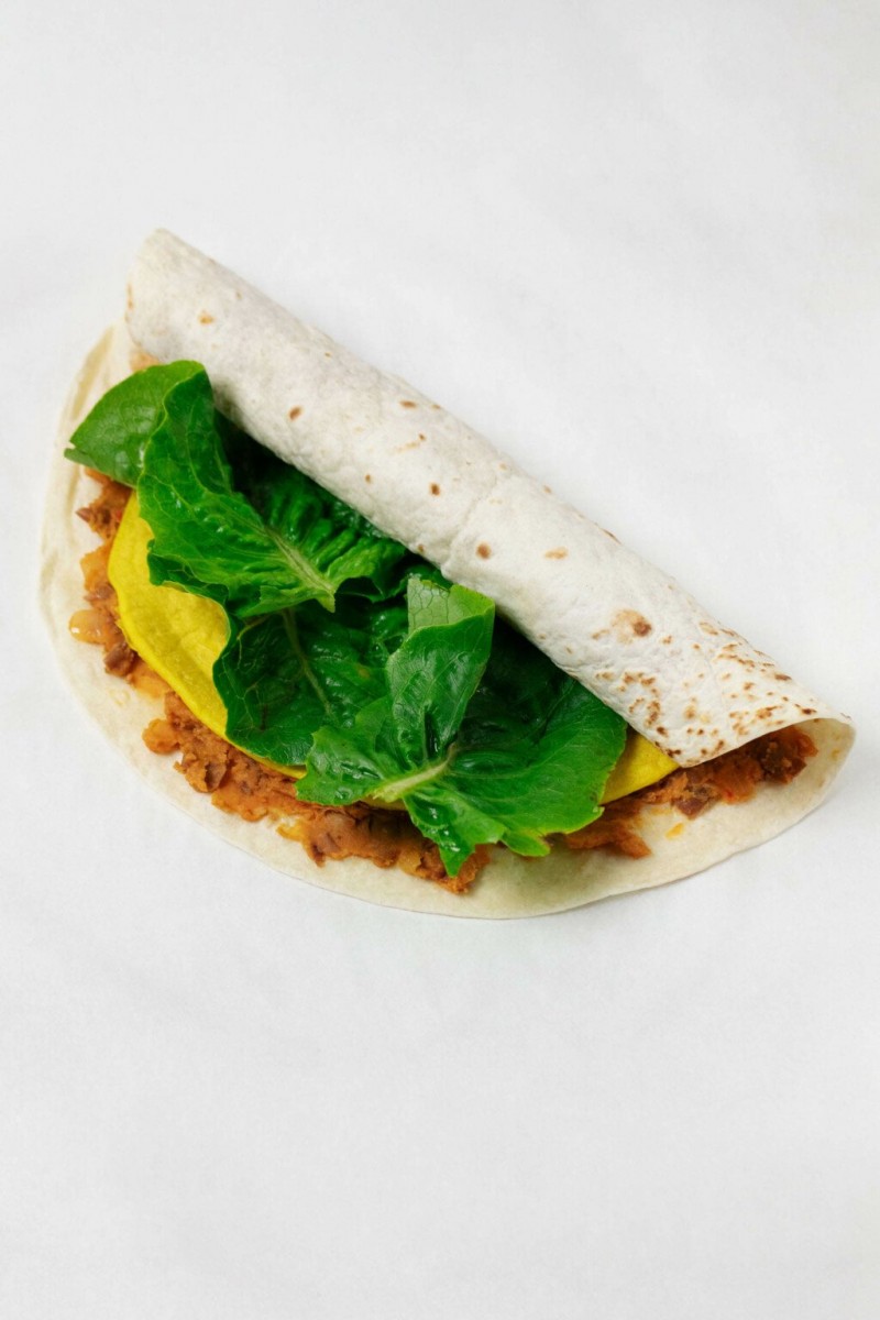 A flour tortilla has been filled and is being rolled up. It's resting on a white work surface.