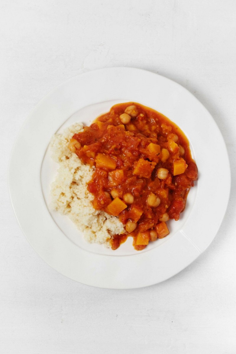 A large, rimmed white plate is covered in couscous and a Moroccan-inspired, butternut chickpea stew.