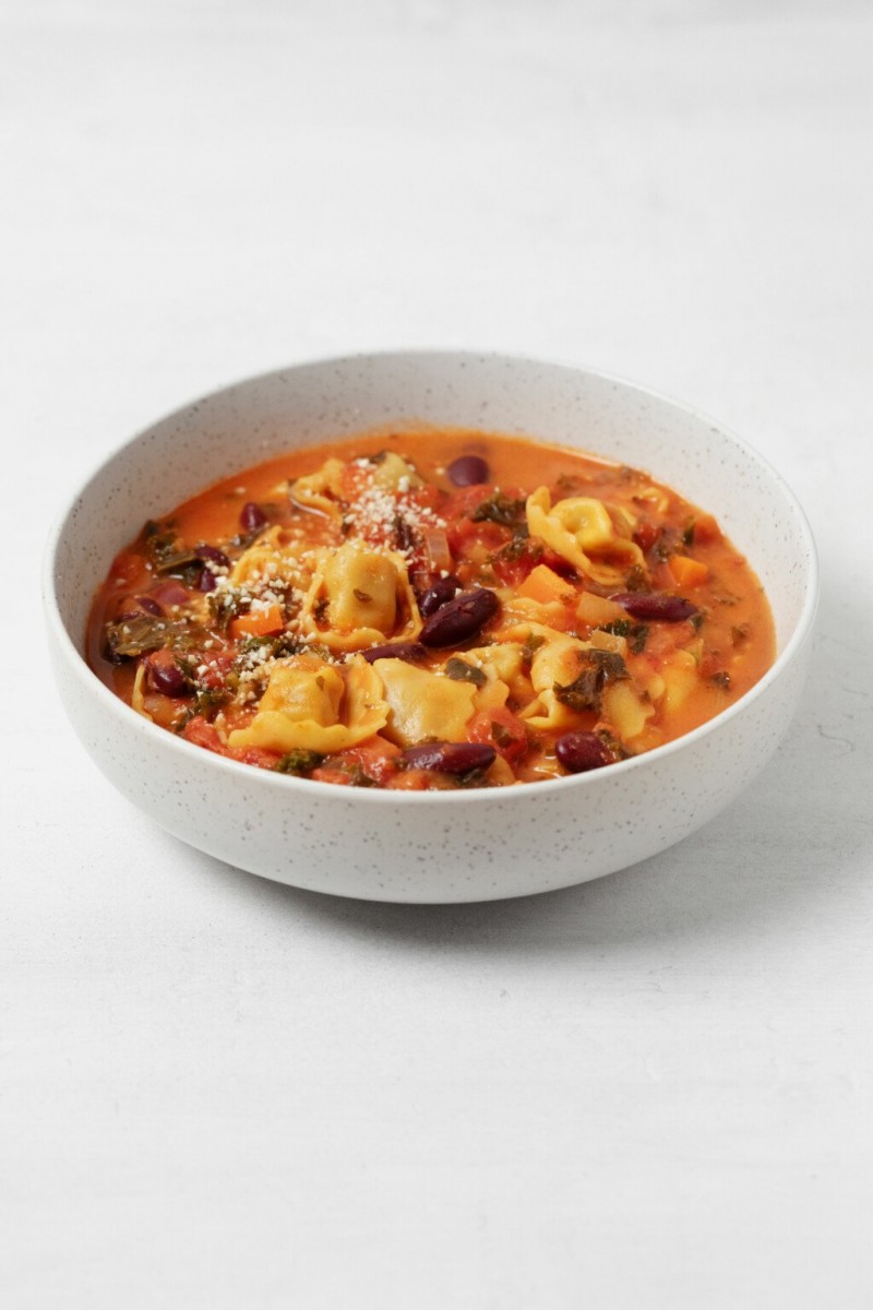 A round, shallow, white bowl of tomato-based vegan soup with pasta and vegetables is resting on a white surface.