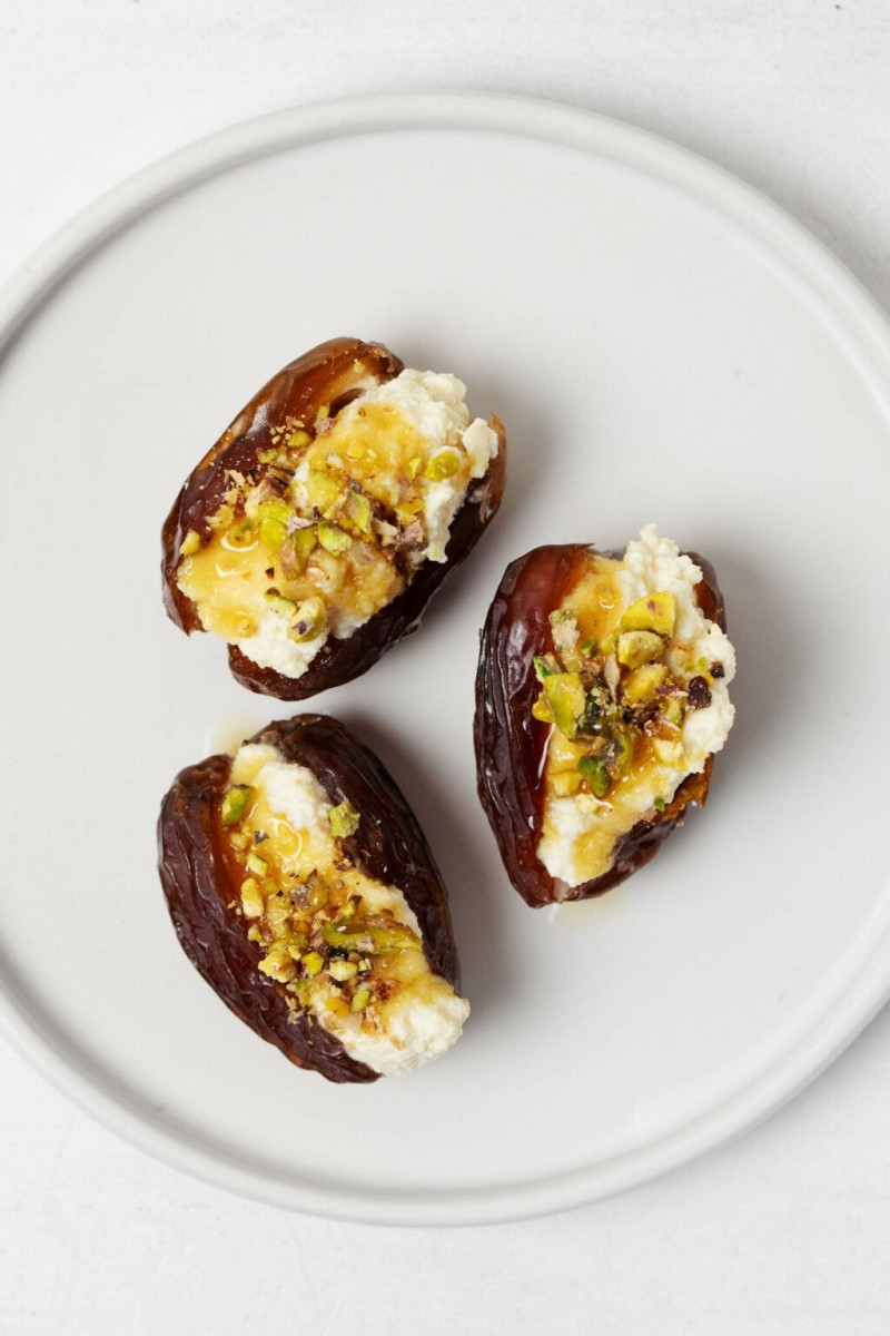 Three sweet and savory stuffed dates, which are topped with pistachio nuts, rest on a white, round appetizer plate.