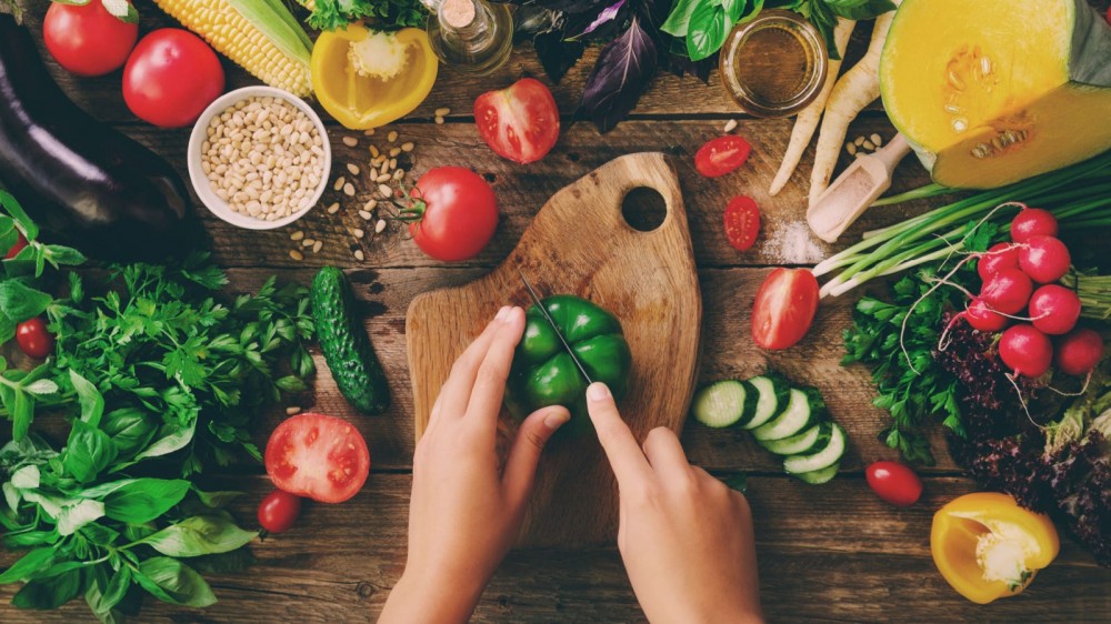 A person chops a bell pepper on a wooden cutting board surrounded by other fruits and vegetables.