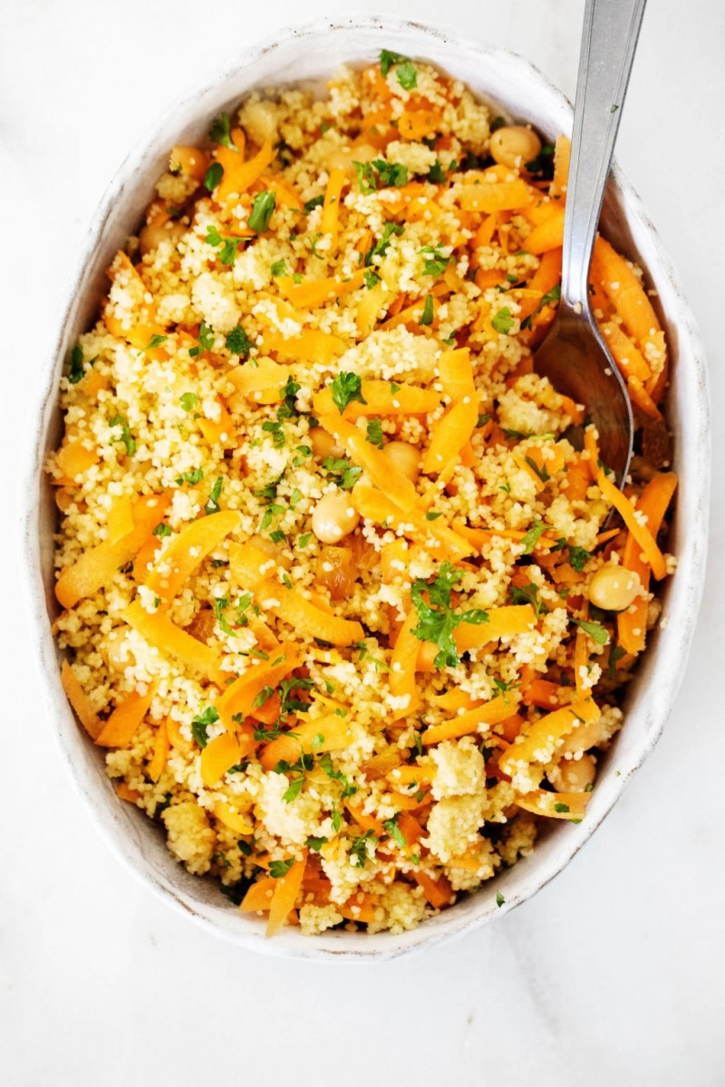 An oval, white ceramic dish is filled with a chickpea couscous salad. It rests on a white surface.