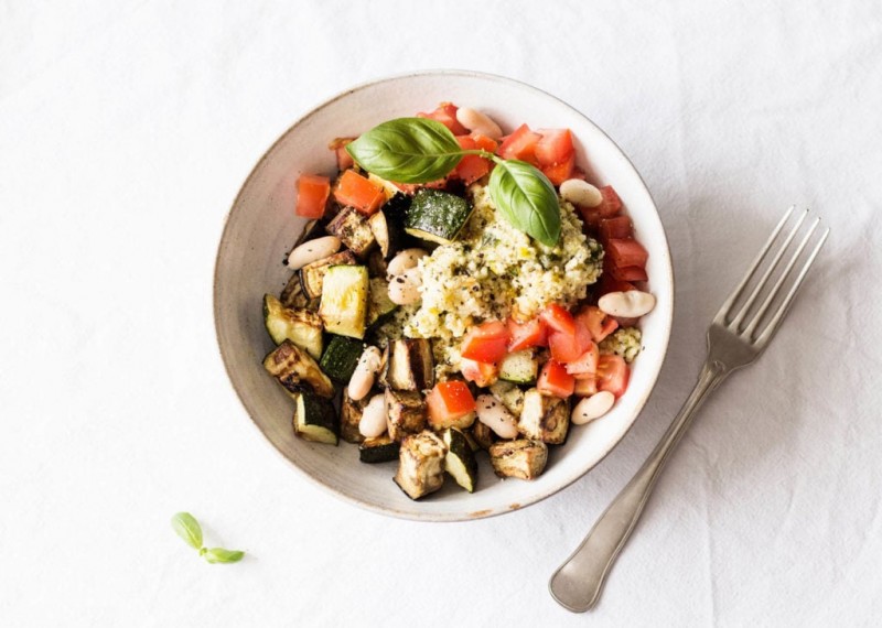 A white bowl has been filled with tomato, zucchini, eggplant, whole grains, and legumes. It rests on a white cloth.