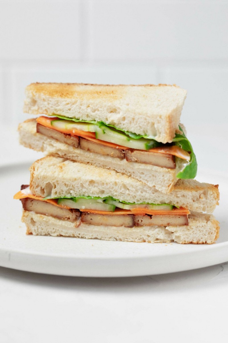 An angled image of a pickled vegetable and smoky tofu sandwich. The sandwich has been cut in half, revealing a cross section with cucumber, carrot, and mayonnaise, along with the tofu. 