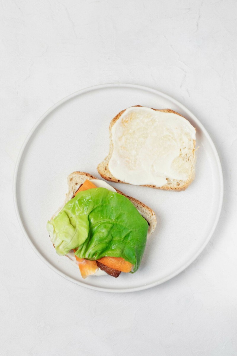 Two slices of toast rest on a round, white plate. One slice is topped with a layer of creamy mayonnaise, the other with butter lettuce.