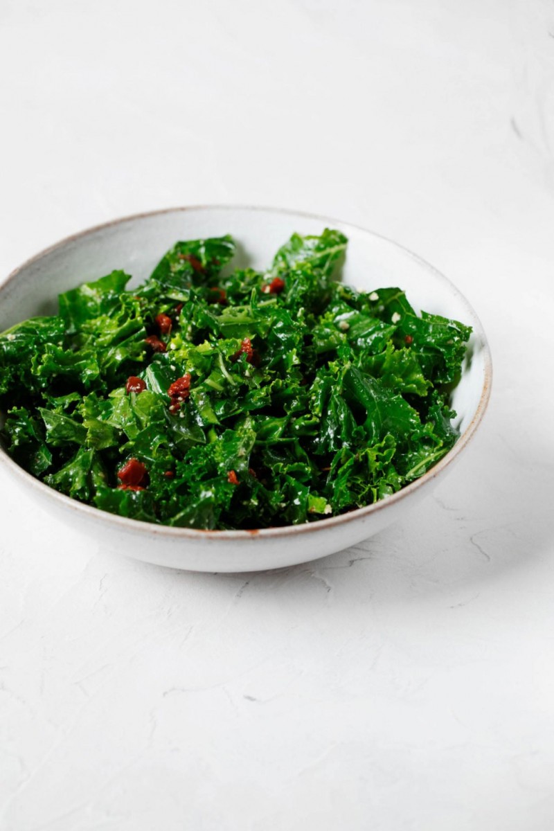 An angled photograph of a bed of dark, leafy greens, which are specked with small pieces of sun-dried tomato. They're sitting in a ceramic bowl.