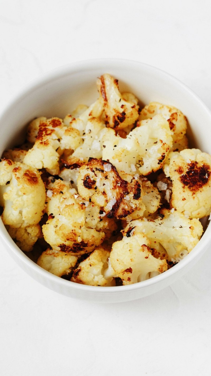 An overhead image of a white bowl, which is filled with roasted frozen cauliflower florets that are topped with flaky sea salt.