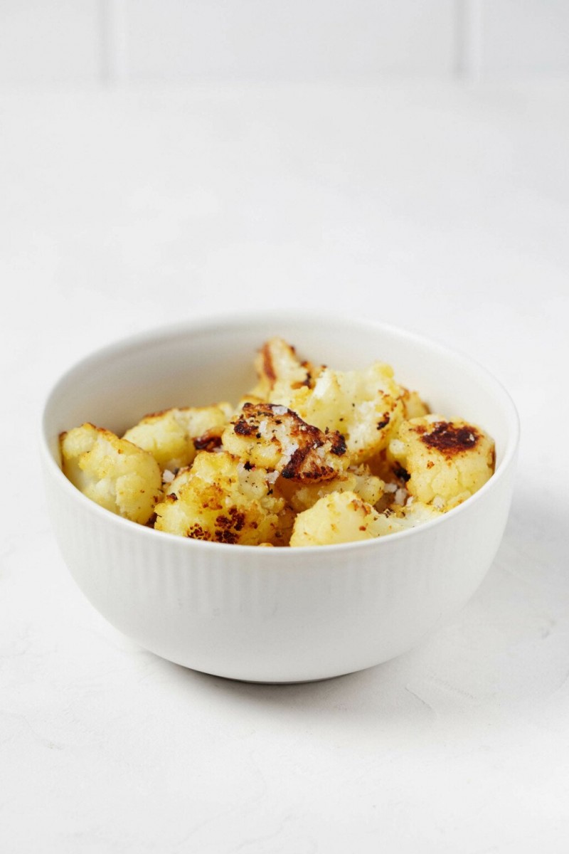 A white, ceramic bowl is resting on a white surface, with a white tile backdrop behind it. The bowl is filled with pieces of roasted cauliflower.