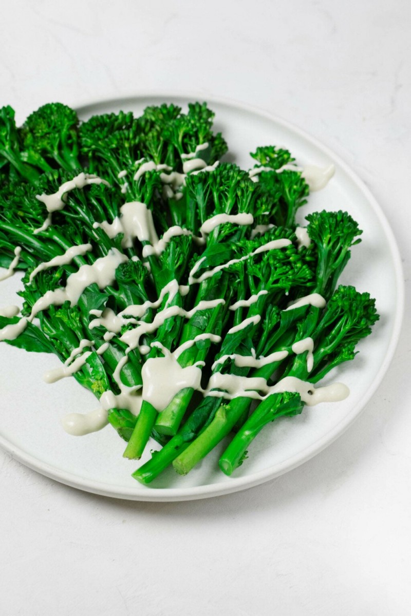 An overhead image of bright green, steamed broccolini resting on a white, round plate. The broccolini is drizzled with a creamy white tahini dressing.