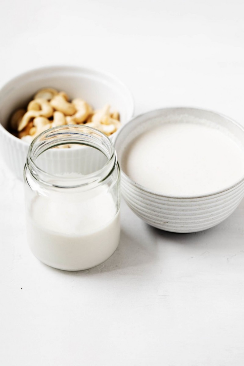 A jar and round bowl have been filled with vegan cashew cream. Another bowl of raw cashews is in the background.