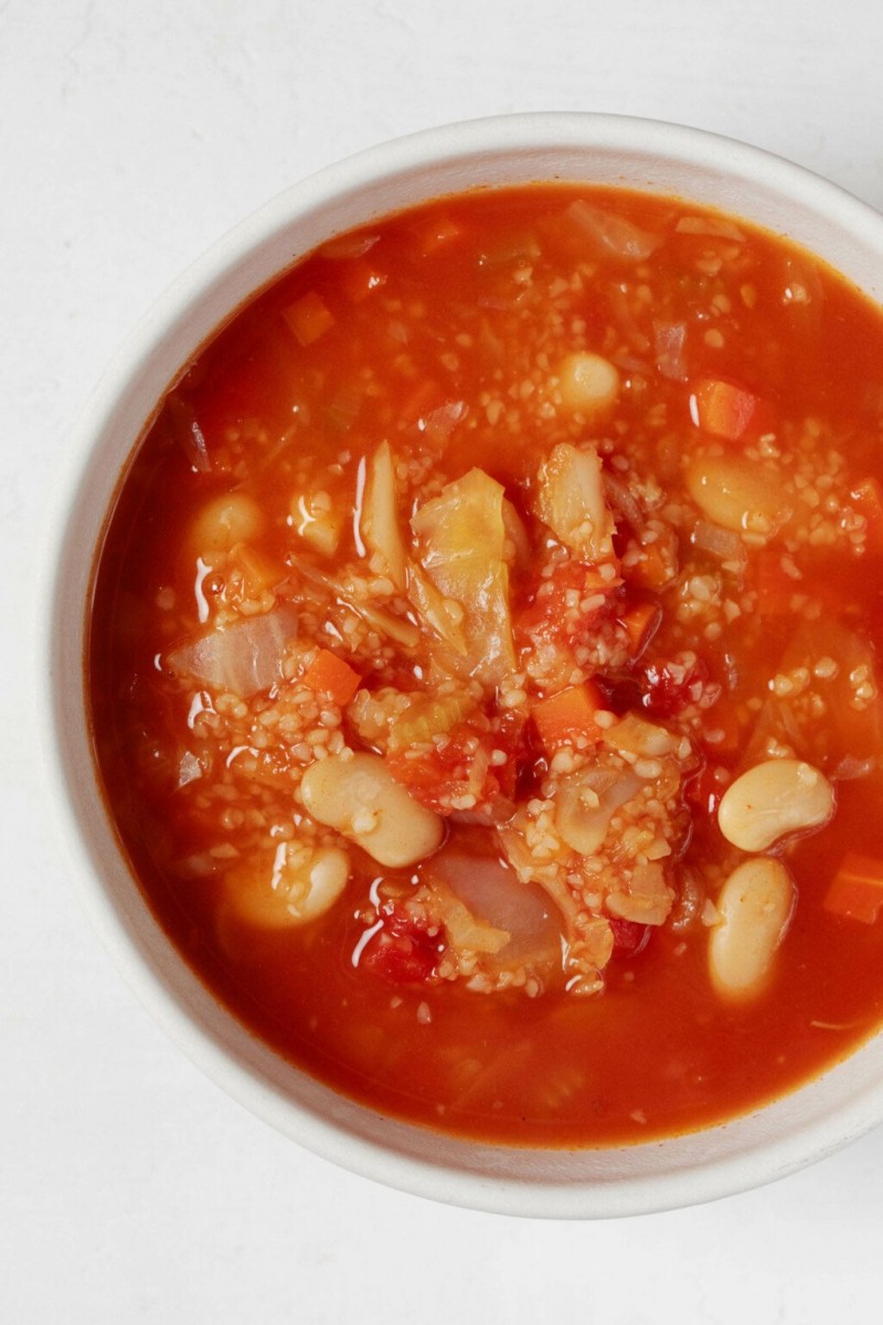 An overhead image of a white bowl of red-colored soup, which is filled with bulgur wheat and white beans.