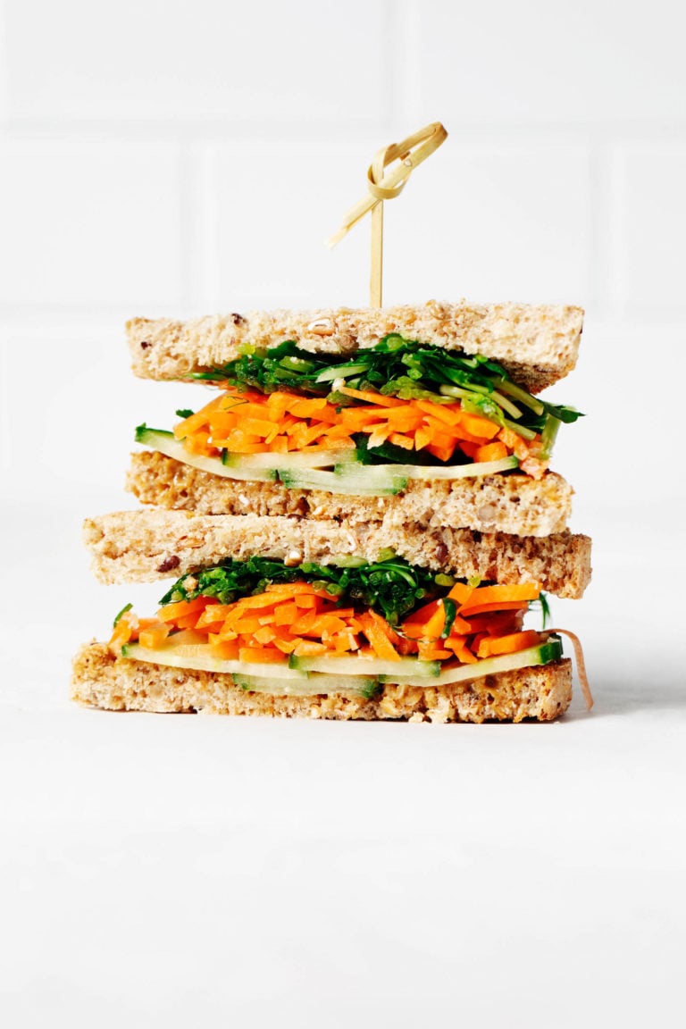 A sandwich has been assembled with layers of carrot, sprouts, and cucumber slices. It's resting on a white surface against a white brick backdrop.